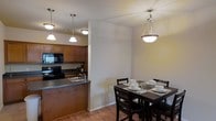 This single-floor residence showcases an open-concept layout that connects the kitchen and dining area into a cohesive living space. The kitchen is equipped with rich, warm-toned wood cabinetry, black appliances including a range and built-in microwave, and a peninsula with a dark countertop that doubles as a workspace and casual gathering spot. A double sink is integrated into the peninsula, adding functional convenience while maintaining an open sightline to the adjacent dining area.

The dining space is defined by an elegant pendant light fixture suspended from the ceiling, complementing the matching pendant lights that illuminate the kitchen. A dark-wood dining table with matching chairs sits beneath this fixture, creating an inviting area for meals. Neutral-toned walls and hardwood-style flooring flow throughout both spaces, tying the design together with warmth and continuity. The light, airy color palette of the walls contrasts pleasantly with the darker furnishings and cabinetry, lending the space a balanced and welcoming ambiance overall.