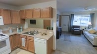 This single-floor residence presents a practical and welcoming layout, with the kitchen and living area forming the heart of the home. The kitchen is equipped with warm oak cabinetry that provides generous upper and lower storage, complemented by light-toned laminate countertops. A double-basin sink sits beneath the upper cabinets, and a white dishwasher is neatly integrated beneath the counter. A mirrored backsplash adds a subtle sense of depth to the kitchen space, while small decorative plants introduce a touch of greenery.

The kitchen opens directly into the living area, which features neutral carpet flooring that extends toward the main entry door. A rug near the entryway defines the transition zone, and a seating area with a cushioned chair is visible in the background. The front door, fitted with a small window panel, allows natural light to filter into the space, enhancing the overall brightness. The overall ambiance is clean, understated, and functional, making it a comfortable everyday living environment.