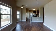 This single-floor space is defined by its open-concept layout and warm, cohesive design. Rich, dark hardwood flooring runs throughout, anchoring the space with a sense of depth and sophistication. The expansive main living area is bathed in natural light streaming through large windows, offering views of the surrounding exterior while keeping the interior bright and welcoming.

The kitchen is a standout feature, elevated slightly by a breakfast bar peninsula that creates a natural boundary between the cooking and living zones without sacrificing the open feel. Dark cabinetry lines the kitchen walls, complemented by stainless steel appliances including a refrigerator, and a sleek countertop surface at the bar. A row of modern pendant lights hangs above the peninsula, providing focused task lighting and adding a stylish decorative element. An additional ceiling fixture illuminates the kitchen workspace.

The neutral, earthy wall tones throughout the space create a calm and inviting ambiance that ties all areas together harmoniously. White baseboards offer a clean contrast against the dark floors. A doorway at the rear hints at additional rooms beyond, and a glass-panel door allows light to pass through, reinforcing the airy, connected atmosphere. Overall, this space balances modern design with practical, comfortable living.