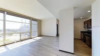 This single-floor space showcases a modern, open-concept layout defined by clean architectural lines and a neutral color palette. The main living area is bathed in natural light, courtesy of large floor-to-ceiling windows fitted with roller shades, which frame views of the surrounding urban landscape. Sunlight streams across the light-toned hardwood-style flooring, enhancing the sense of openness and warmth throughout the space.

The walls are finished in a crisp off-white tone, complemented by warm beige accent walls that add subtle depth without overwhelming the minimalist aesthetic. A structural column near the center of the space serves as a natural divider, subtly delineating distinct functional zones while maintaining the open flow of the layout. Dark baseboards provide a sharp contrast against the lighter flooring, adding a refined, contemporary detail.

Toward the rear of the main area, a doorway leads to an adjoining room, suggesting additional functional space beyond the primary living zone. Electrical outlets and switch plates are neatly positioned throughout, indicating thoughtful placement for everyday use. Overall, the space presents a clean, move-in-ready environment with a bright, airy ambiance well-suited to modern living.