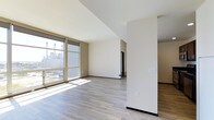 This single-floor space showcases a modern, open-concept layout defined by clean architectural lines and a neutral color palette. The main living area is bathed in natural light, courtesy of large floor-to-ceiling windows fitted with roller shades, which frame views of the surrounding urban landscape. Sunlight streams across the light-toned hardwood-style flooring, enhancing the sense of openness and warmth throughout the space.

The walls are finished in a crisp off-white tone, complemented by warm beige accent walls that add subtle depth without overwhelming the minimalist aesthetic. A structural column near the center of the space serves as a natural divider, subtly delineating distinct functional zones while maintaining the open flow of the layout. Dark baseboards provide a sharp contrast against the lighter flooring, adding a refined, contemporary detail.

Toward the rear of the main area, a doorway leads to an adjoining room, suggesting additional functional space beyond the primary living zone. Electrical outlets and switch plates are neatly positioned throughout, indicating thoughtful placement for everyday use. Overall, the space presents a clean, move-in-ready environment with a bright, airy ambiance well-suited to modern living.