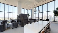 This elevated amenity space is defined by its dramatic architectural design, combining soaring double-height ceilings with expansive floor-to-ceiling curtain-wall glazing that floods the interior with natural light and frames unobstructed panoramic city skyline views. The open-plan layout seamlessly connects multiple functional zones, creating a cohesive and refined atmosphere throughout.

At the heart of the main lounge, a sculptural dark stone fireplace serves as a commanding focal point, clad in polished geometric panels and flanked by plush grey velvet lounge chairs. A contemporary multi-globe chandelier descends from above, complementing the clean white palette of the space. Adjacent seating areas are thoughtfully arranged to maximize the sweeping views beyond the glass.

A conference or meeting area features a bold white marble accent wall housing a mounted display screen, paired with a sleek white island and warm-toned wood and leather seating. A dedicated lounge area opens directly to an outdoor terrace via large black-framed glass doors, offering access to poolside amenities and additional outdoor seating. Neutral carpeting, recessed lighting, and minimalist furnishings throughout reinforce the space's polished, contemporary character.