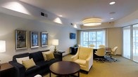 This single-floor space is thoughtfully designed to balance comfort and functionality in a clean, modern aesthetic. The main living area features a deep navy sofa adorned with coordinating accent pillows, paired with a cream upholstered armchair that together create an inviting seating arrangement. A dark-stained round coffee table anchors the space, while a matching side table with a classic table lamp adds warmth and a refined touch.

The room benefits from a combination of recessed lighting and a large, elegant drum pendant light, ensuring the space is well-lit at all times of day. A large multi-pane window along one wall floods the interior with natural light and offers views to the exterior, enhancing the sense of openness.

Adjacent to the lounge area, a dedicated workspace is equipped with a desk and rolling chairs, making it well-suited for productive work sessions. Neutral-toned walls in a soft gray-blue complement the warm carpeted flooring, while framed wall art adds a polished, finished look to the décor. The overall ambiance is calm and professional, blending residential comfort with practical amenities in a cohesive, well-organized layout.