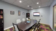 This conference room is a well-appointed meeting space designed to support professional gatherings and collaborative work sessions. At its center sits a dark espresso-toned oval conference table surrounded by eight chairs — a mix of ergonomic mesh-back and upholstered seating — providing comfortable accommodation for small to mid-sized groups. A wall-mounted flat-screen television serves as the room's primary presentation and media display, positioned for clear visibility from all seats.

The room features a drop ceiling with recessed lighting that evenly illuminates the space, complemented by soft gray walls that contribute to a focused, professional atmosphere. Two dark wood bookshelves flank the walls, stocked with reference materials and decorative items, while several framed prints add visual interest without distracting from the room's functional purpose.

A bold, patterned carpet in shades of magenta, olive green, and gold adds a vibrant contrast to the otherwise neutral color palette, lending the space a modern, contemporary feel. Glass partition walls with aluminum framing line one side of the room, offering visual connectivity to adjacent office spaces while maintaining an enclosed, private environment suitable for confidential discussions and focused meetings.