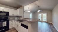 This single-story residence offers a thoughtfully designed layout that blends functionality with comfort. The kitchen is a standout feature, showcasing crisp white shaker cabinetry, granite-style countertops, a peninsula breakfast bar, and a full suite of black appliances including a range, over-the-range microwave, refrigerator, and dishwasher. Wood-look flooring runs throughout the kitchen and extends into the open living and dining areas, which benefit from vaulted ceilings, pendant lighting, and a ceiling fan. French doors along the exterior wall flood the living space with natural light and provide views of the surrounding greenery. A dedicated dining area connects seamlessly to the living room, creating an inviting, open flow. The bedroom features plush carpeting, crown molding, a ceiling fan, and a large window overlooking mature trees. The bathroom is well-equipped with a wide granite-style vanity, white cabinetry, wood-look flooring, and a large mirror with vanity lighting. An adjacent walk-in closet with built-in shelving and hanging rods provides ample storage. Throughout the space, neutral gray tones, white trim, and consistent finishes create a clean, cohesive aesthetic.
