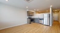 This single-floor unit showcases a clean, contemporary design with light hardwood-style flooring running consistently throughout the main living areas, creating a cohesive and airy feel. The open-concept layout connects the living space directly to the kitchen, maximizing the sense of openness.

The kitchen is well-appointed with a full suite of stainless steel appliances, including a refrigerator, electric range, built-in microwave, and dishwasher. Warm-toned wood cabinetry provides ample storage both above and below the granite countertops, which offer generous prep space. The L-shaped configuration makes efficient use of the kitchen footprint.

The living area is bright and open, benefiting from natural light through windows fitted with blinds. Recessed lighting and a ceiling-mounted HVAC unit provide comfortable climate control throughout the space. The neutral white walls offer a versatile backdrop adaptable to various interior styles.

The bathroom features light tile flooring, a white vanity with granite countertop and undermount sink, and a tub-shower combination with white subway tile surround. The layout is functional and clean, maintaining the modern aesthetic found throughout the unit.
