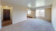 The Almond is a thoughtfully laid-out one-bedroom, one-bathroom residence spread across a single floor. The living area is generously proportioned, featuring neutral-toned walls, plush wall-to-wall carpeting, and large windows that invite abundant natural light while framing pleasant outdoor views of surrounding trees and water. A defined transition leads into a separate dining or flex area with wood-look flooring, providing a seamless connection to the kitchen.

The galley-style kitchen is well-equipped, featuring warm honey-toned wood cabinetry offering ample upper and lower storage, light countertops, a double-basin sink, and white gas appliances including a range and refrigerator. Wood-look flooring extends throughout this area, adding warmth and easy maintenance.

The bedroom is a comfortable retreat with soft carpeting, neutral walls, and a built-in closet with shelving for organized storage. An additional storage closet with built-in shelving is also accessible from the living area, adding practical utility to the floor plan.

The bathroom is clean and functional, featuring a full tub and shower combination with white tile surround, mosaic tile flooring, a vanity with sink, and chrome fixtures that lend a crisp, polished finish to the space.