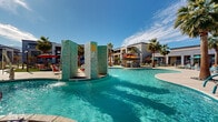 Located at 3875 W Dobbins Rd in Phoenix, AZ, this expansive outdoor amenity area delivers a resort-style experience designed for relaxation and social gathering under the Arizona sun. At the heart of the space is a generously sized freeform swimming pool with crystal-clear turquoise water, complemented by a striking tiled water feature that adds both visual interest and a soothing ambiance. A separate, smaller wading pool provides an additional area for leisure. Surrounding the pools, a broad concrete deck offers ample room for circulation and lounging, lined with vibrant teal and orange chaise lounge chairs and colorful umbrellas that infuse the space with a lively, modern palette. Lush artificial turf lawn areas are interspersed throughout, providing comfortable spots for sunbathing and lawn games such as cornhole. A covered outdoor bar and lounge area with barstool seating and a mounted television serves as a social hub. Private cabanas with sheer curtains offer shaded retreats. Towering palm trees frame the entire courtyard, enhancing the tropical atmosphere. The surrounding multi-story residential buildings with balconies provide a sense of enclosure while the open sky above ensures abundant natural light throughout the day.