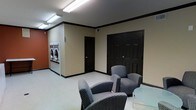 This single-floor communal facility combines a functional laundry room with a relaxing lounge area, making it a practical and comfortable space for residents or guests. The interior features neutral beige walls accented by bold black crown molding and baseboards, lending the space a clean, polished look. Overhead fluorescent lighting ensures the room is bright and well-illuminated throughout.

The laundry area is equipped with stacked front-loading washers and dryers, neatly arranged along one wall and easily accessible. Adjacent to the laundry equipment, the lounge area provides a welcoming place to wait, furnished with cushioned armchairs featuring a subtle geometric pattern and a small side table, encouraging a comfortable and relaxed atmosphere.

Multiple dark-paneled doors with classic raised molding lead to additional rooms or utility areas, contributing to the space's organized layout. The light-toned flooring ties the room together, maintaining a cohesive and tidy appearance throughout. Overall, this well-appointed facility balances utility and comfort, offering everything needed for a convenient and pleasant shared laundry experience.