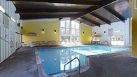This indoor aquatic facility is designed to offer a comfortable and inviting swimming environment year-round. The centerpiece is a generously sized rectangular swimming pool with clear, blue water, accessible via stainless steel entry rails and marked with depth indicators for swimmer safety. The architectural design is defined by a striking vaulted ceiling with dark wood beam detailing, lending the space a warm yet open character. Large, floor-to-ceiling windows along the far wall invite abundant natural light, framing outdoor views of surrounding greenery and creating a bright, airy atmosphere throughout the day. The interior walls are finished in a warm yellow tone, enhancing the sense of warmth and energy within the space. The pool deck is lined with lounge chairs and patio-style seating arranged beneath umbrella stands, providing ample space for relaxation before or after a swim. Wall-mounted sconces add soft ambient lighting to complement the natural light. On one side, a dedicated seating area with additional furniture offers a comfortable spot to gather. The overall ambiance is clean, functional, and welcoming, making this facility well-suited for both recreational swimming and leisure.