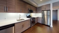 This contemporary single-floor residence offers a well-appointed layout designed with modern finishes throughout. The kitchen serves as a standout feature, showcasing floor-to-ceiling wood-grain cabinetry in a rich walnut tone, a crisp white quartz countertop, and a white subway tile backsplash illuminated by under-cabinet lighting. A full suite of stainless steel appliances — including a gas range, built-in microwave, dishwasher, and refrigerator — completes this functional and stylish culinary space. Dark hardwood flooring runs throughout the main living and kitchen areas, lending warmth and a cohesive feel to the open layout. The bedroom features soft, light gray carpeting, clean white walls, and a sliding double-door closet with sleek hardware, providing generous storage. A dedicated storage or utility nook is also present, featuring neutral walls and dark wood-toned door accents. The bathroom is finished with bright white square tile surrounding a full soaking tub and shower combination, complemented by brushed bronze fixtures for an elevated touch. Light-toned floor tiles and clean white surfaces contribute to a fresh, minimalist aesthetic throughout the bathroom. Overall, the space presents a cohesive modern design with quality finishes and practical functionality.