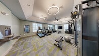 This fitness center offers a comprehensive workout environment designed with both functionality and aesthetics in mind. The space is finished with soft sage-green walls and distinctive patterned carpet tiles in shades of gray and yellow-green, creating a fresh, energizing atmosphere throughout.

The cardio zone features multiple treadmills and an elliptical machine positioned along windowed walls, allowing natural light to fill the area and providing exterior views during workouts. A ceiling fan and circular ring-pendant light fixtures illuminate the space stylishly while maintaining comfortable airflow.

The strength training area is well-stocked with a full dumbbell rack, adjustable weight bench, stability ball, a multi-station cable machine, and a heavy punching bag. A wall-mounted flat-screen television serves the strength zone, while a smart mirror fitness display is mounted near the entrance, offering interactive workout guidance.

Safety and convenience are addressed with a mounted AED unit and a dual-function water fountain with bottle-filling station. The entry corridor features elegant herringbone-patterned hardwood flooring and frosted glass doors, providing a polished transition into the fitness area. The raised ceiling with tray detailing adds a sense of volume and sophistication to the overall space.