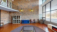 Located at 3020 Glencrest Road in Burlington, Ontario, this distinctive living space makes a bold architectural statement from the moment you enter. The room is defined by its impressive double-height ceilings, which lend a grand, open sense of volume rarely found in a single-floor layout. Warm terracotta tiles laid in a classic herringbone pattern span the entire floor, anchoring the space with rich, earthy tones.

A full-height curtain wall of steel-framed windows dominates one side of the room, flooding the interior with abundant natural light and offering clear views of the surrounding neighbourhood streetscape. Anchoring the window wall is a sleek, modern electric fireplace unit, providing a striking contrast of contemporary comfort against the room's more traditional elements.

Exposed brick walls add texture and warmth to the space, accented by vintage-style wall lanterns that evoke a classic charm. An ornate wrought-iron lattice partition with a decorative arch serves as an elegant interior divider, while a curved mezzanine balcony overhead features intricate ironwork railings, reinforcing the space's sophisticated character.

The living area is furnished with leather sofas and paired lounge chairs, arranged around a central circular area rug. A modular open-shelf unit and a vibrant blue sideboard complete the functional yet characterful design of this remarkable communal space.
