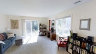 This two-story home offers two bedrooms and one and a half bathrooms across a well-utilized floor plan. The main living area showcases abundant natural light streaming through large windows and a sliding glass door that opens directly to an outdoor patio, creating a seamless indoor-outdoor connection. The carpeted room features neutral walls and a clean, open feel that lends itself to a variety of uses, whether as a living space, home office, or both. The sliding door frames views of a lush backyard with mature greenery, enhancing the sense of openness. Multiple windows positioned along the walls allow sunlight to flood the interior throughout the day, keeping the atmosphere bright and airy. Built-in shelving opportunities and ample floor space provide flexibility for furniture arrangement and storage. The overall ambiance is calm and welcoming, with a light color palette that makes the space feel larger and more open. The two-story layout offers a natural separation between living and sleeping areas, contributing to a functional and comfortable daily living experience.