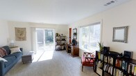 This two-story home offers two bedrooms and one and a half bathrooms across a well-utilized floor plan. The main living area showcases abundant natural light streaming through large windows and a sliding glass door that opens directly to an outdoor patio, creating a seamless indoor-outdoor connection. The carpeted room features neutral walls and a clean, open feel that lends itself to a variety of uses, whether as a living space, home office, or both. The sliding door frames views of a lush backyard with mature greenery, enhancing the sense of openness. Multiple windows positioned along the walls allow sunlight to flood the interior throughout the day, keeping the atmosphere bright and airy. Built-in shelving opportunities and ample floor space provide flexibility for furniture arrangement and storage. The overall ambiance is calm and welcoming, with a light color palette that makes the space feel larger and more open. The two-story layout offers a natural separation between living and sleeping areas, contributing to a functional and comfortable daily living experience.