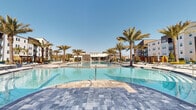 This outdoor amenity space is designed with a resort-inspired aesthetic, centered around a generously sized, free-form swimming pool with exceptionally clear, blue water. The pool's curvilinear shape includes a gradual entry with stepped tiers, making it both visually striking and highly accessible. The surrounding pool deck is finished in large-format, multi-toned pavers in cool grey and blue hues, lending a sophisticated, contemporary look throughout the space.

Tall palm trees are strategically placed around the perimeter, providing a tropical atmosphere while offering natural shade across the deck. Modern lounge chairs with teal accent cushions and retractable umbrellas are arranged along the poolside, creating comfortable areas for sunbathing and relaxation. Slatted wooden privacy screens and manicured landscaping with mulched planting beds define the boundaries of the space, adding warmth and texture.

At the rear of the pool area, an open-air pavilion with a gabled roof, board-and-batten siding, and large open archways serves as a communal gathering space. A decorative circular pendant fixture accents the entrance, while ceiling fans and iron-gated entries enhance the structure's inviting character. The overall ambiance is bright, open, and polished, ideal for outdoor leisure.