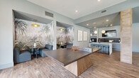 Located at 2555 Cobb Pkwy in Kennesaw, Georgia, this single-floor amenity space offers a well-appointed, open-concept layout designed for both relaxation and social gatherings. Wood-look tile flooring runs throughout, providing a warm, cohesive foundation that ties each zone together seamlessly.

The expansive living area is anchored by a large, plush sectional sofa, accent chairs, and layered area rugs, all bathed in natural light from floor-to-ceiling windows with outdoor views and elegant draping curtains. A sculptural multi-arm chandelier adds a contemporary focal point overhead.

Adjacent to the living area, a dedicated dining zone features a wooden trestle-style dining table surrounded by upholstered seating, illuminated by a distinctive woven pendant light. Large windows flood this corner with natural light, while curated wall art and a credenza add refined detail.

The game and lounge area showcases a wood-paneled tray ceiling accent, a foosball table, and a built-in bar counter with bar stools, a mounted TV, and a striking tropical mural that energizes the space. Structural columns with textured stone-like cladding serve as elegant architectural dividers throughout, adding character and defining each functional zone within the open floor plan.