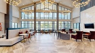 Located at Integra Village in Daytona Beach, Florida, this elegantly designed single-floor clubhouse lounge offers a refined and welcoming atmosphere across a generously sized open floor plan. The space makes an immediate impression with its dramatic vaulted ceiling, which soars overhead and is adorned with a series of sculptural ring chandeliers featuring warm globe lighting. Floor-to-ceiling black-framed windows line the perimeter, flooding the interior with abundant natural light and providing sweeping outdoor views of the surrounding landscape and palm trees.

The entry foyer features large-format stone tile flooring, a wood console with a digital display, and clerestory windows that maintain the airy, open feel from the moment of arrival. The living area is anchored by a striking floor-to-ceiling marble fireplace wall with an embedded television, framed by a slate-blue accent panel — a clear focal point of the room. Rich burgundy velvet armchairs and curved upholstered seating are thoughtfully arranged throughout the space.

The dining area offers built-in booth-style banquette seating paired with marble-top bistro tables and warm leather chairs. A game room is visible in the background, adding a recreational dimension to the space. Large glass partition walls with dark steel framing define areas while maintaining visual connectivity, contributing to the overall sense of openness and modern sophistication.
