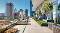 This expansive rooftop terrace occupies the upper level of a modern high-rise building, offering a thoughtfully designed outdoor environment that blends urban elegance with lush greenery. The terrace is finished with large-format, light-toned stone pavers that extend across the entire deck, creating a clean and refined aesthetic throughout.

Landscaping is a defining feature of the space, with mature trees planted in substantial raised planters positioned along the building's floor-to-ceiling glass curtain wall facade. Neatly trimmed hedges and low ground cover plantings fill the planters, introducing vibrant greenery that contrasts beautifully against the contemporary architecture. The glass facade reflects the sky and surrounding cityscape, adding depth and visual interest to the setting.

Multiple seating areas are distributed across the terrace, furnished with woven lounge chairs, sectional sofas, and accent tables — all designed for outdoor comfort and social gathering. Clear glass balustrades line the perimeter, preserving unobstructed, sweeping panoramic views of the city skyline in multiple directions, including dense downtown towers and broader urban neighborhoods stretching to the horizon.

The terrace benefits from abundant natural sunlight throughout the day. Covered corridor sections along the building edge offer shaded retreats, while open zones invite full sun exposure. The overall ambiance is one of elevated urban sophistication — a premier outdoor amenity space ideal for both relaxation and professional or social gatherings.