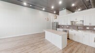 Located at 94 E 11th St in Sioux Falls, South Dakota, this single-floor unit showcases a striking blend of industrial and contemporary design elements throughout. Soaring ceilings with exposed ductwork, reflective tile ceiling panels, and recessed track lighting define the open-concept living and kitchen area, creating a loft-like atmosphere with considerable vertical volume.

The kitchen is a standout feature, equipped with stainless steel appliances including a refrigerator, stove, and dishwasher. Crisp white shaker cabinetry pairs with a neutral tile backsplash, while a central island with pendant lighting provides additional prep and gathering space. Light-toned luxury vinyl plank flooring extends throughout the main living area, enhancing the bright, airy feel.

Multiple carpeted rooms offer flexible use as bedrooms or private spaces, each featuring large windows fitted with roller shades that provide views of the surrounding Sioux Falls cityscape and abundant natural light. Exposed structural elements and ductwork lend an urban industrial character to these rooms as well.

The bathroom is finished to a high standard, featuring large-format marble-look wall tiles in a walk-in shower with a mosaic tile floor inset and a built-in niche. A white vanity with brushed nickel hardware and warm-toned floor tiles complete the clean, polished look. An adjacent walk-in closet with wire shelving provides ample storage.