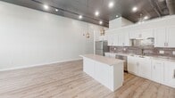 Located at 94 E 11th St in Sioux Falls, South Dakota, this single-floor unit showcases a striking blend of industrial and contemporary design elements throughout. Soaring ceilings with exposed ductwork, reflective tile ceiling panels, and recessed track lighting define the open-concept living and kitchen area, creating a loft-like atmosphere with considerable vertical volume.

The kitchen is a standout feature, equipped with stainless steel appliances including a refrigerator, stove, and dishwasher. Crisp white shaker cabinetry pairs with a neutral tile backsplash, while a central island with pendant lighting provides additional prep and gathering space. Light-toned luxury vinyl plank flooring extends throughout the main living area, enhancing the bright, airy feel.

Multiple carpeted rooms offer flexible use as bedrooms or private spaces, each featuring large windows fitted with roller shades that provide views of the surrounding Sioux Falls cityscape and abundant natural light. Exposed structural elements and ductwork lend an urban industrial character to these rooms as well.

The bathroom is finished to a high standard, featuring large-format marble-look wall tiles in a walk-in shower with a mosaic tile floor inset and a built-in niche. A white vanity with brushed nickel hardware and warm-toned floor tiles complete the clean, polished look. An adjacent walk-in closet with wire shelving provides ample storage.