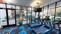 This fitness center combines modern design with comprehensive workout functionality. The space is defined by a striking floor-to-ceiling glass wall that floods the cardio zone with natural light while offering direct views of a landscaped outdoor courtyard, complete with mature trees and a water feature. True-brand treadmills and ellipticals are positioned to take full advantage of this scenic outlook, with mounted flat-screen TVs overhead for entertainment during workouts.

The interior transitions into a well-appointed strength training area featuring a full rack of dumbbells, cable machines, and multi-use weight equipment. Large wall-to-wall mirrors visually expand the space and allow users to monitor form throughout their sessions. Overhead, sleek black ceiling fans circulate air throughout the workout area, complementing the modern industrial aesthetic.

The entrance area features a teal accent wall with an octagonal mirror and a dedicated stretching and functional training zone. Textured wood-panel wall details and clerestory windows add warmth and dimension to the space.

Throughout the facility, a bold geometric-patterned carpet in shades of blue, teal, and gray anchors the contemporary design, tying together the various workout zones into a cohesive, energetic environment.