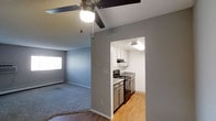 This single-floor space features a practical and straightforward layout with clearly defined living and kitchen areas. The living room is finished with soft blue-gray carpeting and painted in a cool gray tone, creating a calm, neutral atmosphere. A ceiling fan with an integrated light fixture provides both air circulation and overhead lighting, while a baseboard heater offers supplemental warmth. A window allows natural light to filter into the space.

Adjacent to the living area, the kitchen is accessible through a dedicated opening in the wall, giving it a semi-enclosed feel while maintaining visual connection to the main living space. The kitchen is outfitted with white upper cabinetry, gray lower cabinets, and light-toned countertops. Stainless steel appliances, including a gas range and dishwasher, are neatly integrated into the layout. The kitchen floor features warm hardwood-style flooring, contrasting with the carpeted living area. Overhead lighting keeps the kitchen well-illuminated. Overall, the space presents a clean, move-in-ready environment with a cohesive neutral color palette throughout.