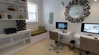 This single-floor home office is thoughtfully designed to balance functionality with contemporary style. The room is finished in crisp white throughout, creating a clean, bright atmosphere enhanced by a window that allows natural light to fill the space. Built-in open shelving lines one wall, providing ample storage and display space while maintaining a streamlined, uncluttered look. Beneath the window sits a built-in bench with a cushioned olive-green seat and coordinating accent pillow, offering a comfortable reading nook or secondary seating area. A minimalist floating desk spans one wall, paired with a modern ergonomic chair on casters, forming a dedicated and efficient workstation. Above the desk, a decorative starburst mirror adds visual interest and dimension to the wall. A sculptural pendant light hangs from the ceiling, contributing both ambient lighting and a design focal point. Light-toned flooring and a neutral area rug ground the space, while carefully chosen accessories—including decorative objects on the shelves and a wall-mounted art panel—add personality without overwhelming the room's clean aesthetic.
