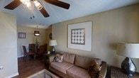 This single-floor space offers a welcoming and cohesive interior designed for comfortable everyday living. The living area serves as the centerpiece of the home, featuring a wicker-framed sofa adorned with decorative floral pillows and a coordinating wicker accent chair. A large framed geometric artwork anchors the main wall, adding a modern touch to the otherwise warm, earthy color palette. A textured ceiling with a ceiling fan overhead provides both visual interest and practical air circulation.

The space flows naturally into a dining area, visible in the background, where dark wood chairs surround a dining table beneath a pendant light fixture with a warm, amber glow. A sunburst wall mirror and a decorative potted plant add character and charm to the transition between spaces. Rich hardwood flooring runs throughout, grounding the design and lending a sense of continuity and warmth across the open layout.

Ambient lighting is thoughtfully layered, with a table lamp providing soft task lighting alongside the overhead fixtures. The overall ambiance is casual yet polished, blending natural textures like wicker and wood with carefully selected decorative accents to create a space that feels both lived-in and well-appointed.