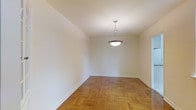 This single-floor space presents a clean and classic aesthetic, characterized by its warm parquet hardwood flooring laid in a traditional basket-weave pattern that adds visual texture and timeless charm to the interior. The walls are finished in a crisp, neutral white, creating a bright and airy atmosphere that serves as a versatile backdrop for a variety of furnishing styles. A pendant light fixture with a bowl-shaped shade hangs from the ceiling, providing focused overhead illumination and serving as a subtle decorative focal point in the main living area. The ceiling height contributes to an open, unencumbered feel throughout the space. Classic white baseboards trim the perimeter of the room, complementing the overall clean and well-maintained character of the interior. The layout appears straightforward and functional, allowing for flexible furniture arrangement. The combination of the warm-toned hardwood flooring and the bright white walls creates a balanced contrast that enhances the sense of space and light. Overall, the space offers a neutral and inviting foundation, well-suited for customization to suit a range of personal styles and functional needs.