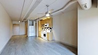 This single-floor urban unit offers a thoughtfully designed open-plan layout that combines living and kitchen areas into one cohesive space. The interior is characterized by an industrial aesthetic, with exposed ceiling pipes and ductwork running alongside a track lighting system, adding architectural interest overhead. A ceiling fan provides both function and style, complementing the loft-inspired design. The kitchen is well-appointed with warm wood cabinetry, a subway-style tile backsplash in a bold geometric pattern, white appliances including a gas range, microwave, and full-size refrigerator. Wide-plank light hardwood flooring flows throughout, lending warmth and continuity to the space. A corridor connects the main living area to additional rooms, offering practical separation within the open concept. The space is further enhanced by a balcony accessed from the main living area, offering views of a shared courtyard below, complete with lounge seating and greenery, as well as the surrounding urban landscape. The overall ambiance blends industrial character with modern comfort, making it a distinctive and functional urban living space.