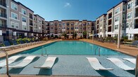 Located at 9015 Eager Rd, this thoughtfully designed outdoor amenity space serves as the centerpiece of a modern multi-story residential community. At its heart is a generously sized resort-style swimming pool with crystal-clear blue water, framed by a broad sun deck outfitted with rows of sleek lounge chairs and shaded umbrella stations. In-pool lounge chairs invite residents to relax at the water's edge, while private canopied cabanas with plush navy-blue cushions provide a more secluded retreat under the open sky.

Surrounding the pool, manicured landscaping features ornamental grasses, flowering plants, and neatly trimmed shrubs that soften the hardscape and add vibrant color throughout the seasons. A dedicated recreation lawn offers artificial turf space ideal for outdoor games such as Jenga, complemented by casual seating and planters with bold, colorful foliage.

The main building entry is marked by a covered pergola-style entrance with clean architectural lines, tile columns, and a welcoming courtyard approach. The overall design of the space balances contemporary aesthetics with functional outdoor living, creating a cohesive environment that encourages relaxation and socializing beneath the open sky.
