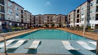 Located at 9015 Eager Rd, this thoughtfully designed outdoor amenity space serves as the centerpiece of a modern multi-story residential community. At its heart is a generously sized resort-style swimming pool with crystal-clear blue water, framed by a broad sun deck outfitted with rows of sleek lounge chairs and shaded umbrella stations. In-pool lounge chairs invite residents to relax at the water's edge, while private canopied cabanas with plush navy-blue cushions provide a more secluded retreat under the open sky.

Surrounding the pool, manicured landscaping features ornamental grasses, flowering plants, and neatly trimmed shrubs that soften the hardscape and add vibrant color throughout the seasons. A dedicated recreation lawn offers artificial turf space ideal for outdoor games such as Jenga, complemented by casual seating and planters with bold, colorful foliage.

The main building entry is marked by a covered pergola-style entrance with clean architectural lines, tile columns, and a welcoming courtyard approach. The overall design of the space balances contemporary aesthetics with functional outdoor living, creating a cohesive environment that encourages relaxation and socializing beneath the open sky.