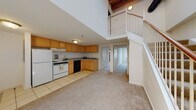 Located on West Broad Street in Richmond, Virginia, this two-floor space blends historic character with practical design. The main level opens into a generous living area with plush carpeting and soaring double-height ceilings that create an impressive sense of volume and openness. The kitchen features warm honey-toned cabinetry arranged in an L-shape, complemented by tile flooring, ample counter space, and a full suite of appliances including a range, dishwasher, and refrigerator. A standout feature of the space is the exposed red brick accent wall, adding a rich architectural detail that speaks to the building's historic roots. Natural light filters in through arched windows framed by the original brick surrounds, further enhancing the character of the interior. Two full bathrooms are appointed with tile flooring, wood-toned vanities with granite-style countertops, and combination tub/shower units. A staircase with a classic white balustrade and warm wood handrail connects the main level to the upper floor, where a carpeted loft overlooks the living area below. The upper level also features exposed wooden beam ceilings, reinforcing the industrial-meets-historic aesthetic throughout. This thoughtfully designed space offers a compelling mix of original architectural charm and functional living areas in the heart of Richmond.
