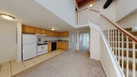 Located on West Broad Street in Richmond, Virginia, this two-floor space blends historic character with practical design. The main level opens into a generous living area with plush carpeting and soaring double-height ceilings that create an impressive sense of volume and openness. The kitchen features warm honey-toned cabinetry arranged in an L-shape, complemented by tile flooring, ample counter space, and a full suite of appliances including a range, dishwasher, and refrigerator. A standout feature of the space is the exposed red brick accent wall, adding a rich architectural detail that speaks to the building's historic roots. Natural light filters in through arched windows framed by the original brick surrounds, further enhancing the character of the interior. Two full bathrooms are appointed with tile flooring, wood-toned vanities with granite-style countertops, and combination tub/shower units. A staircase with a classic white balustrade and warm wood handrail connects the main level to the upper floor, where a carpeted loft overlooks the living area below. The upper level also features exposed wooden beam ceilings, reinforcing the industrial-meets-historic aesthetic throughout. This thoughtfully designed space offers a compelling mix of original architectural charm and functional living areas in the heart of Richmond.