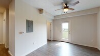 This single-floor unit offers a clean, modern layout with wood-style flooring flowing consistently throughout the space. The living area is bright and open, anchored by a ceiling fan with a light fixture and framed by neutral walls. A glass-paned door leads to a private balcony, allowing natural light to fill the room while offering views of the surrounding landscape. An electrical panel is visibly accessible on one wall, and the entry features a solid dark-colored front door.

The kitchen is a functional highlight of the unit, featuring rich, warm-toned wood cabinetry extending to the upper walls for generous storage. Granite-style countertops provide ample prep space, complemented by a full suite of stainless steel appliances including a range, microwave, dishwasher, and refrigerator. Recessed lighting above keeps the kitchen well-lit.

The bathroom is finished in a clean, neutral palette, featuring a single-sink vanity with a white countertop, wood-toned cabinetry beneath, a large mirror, and a full tub-shower combination with built-in shelving. Wood-style flooring continues into this space, maintaining a cohesive aesthetic throughout the unit.