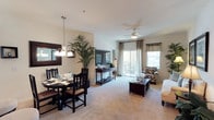 This single-floor residence is part of a well-maintained Mediterranean-style community, characterized by warm stucco exteriors, red-tiled rooflines, and covered parking. The building's architecture reflects a cohesive, southwestern-inspired design set under bright, sunny skies.

Inside, the open-concept layout connects the dining and living areas into one inviting, light-filled space. The dining area features a dark wood table and chairs set beneath an elegant pendant chandelier, complemented by a large decorative mirror that enhances the sense of depth. A tall indoor palm plant adds a natural, organic touch to the interior.

The living area extends seamlessly from the dining space, finished with plush carpeting and neutral wall tones that create a warm and comfortable ambiance. A ceiling fan with a light fixture serves as a functional centerpiece overhead. A console sideboard adds additional storage and display space between the two zones.

Sliding glass doors lead to a private balcony, allowing natural light to pour into the living space while offering views of the surrounding community. The balcony provides an inviting outdoor retreat overlooking the tree-lined courtyard and neighboring buildings. Draped curtains with decorative valances frame the windows and glass doors, adding a polished, finished look to the overall interior design.
