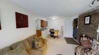 This single-floor unit offers an efficient, open-concept layout that seamlessly connects the living, dining, and kitchen areas into one cohesive space. The living area is anchored by plush carpeting and features a comfortable seating arrangement accented with decorative throw pillows, creating a warm and inviting atmosphere. A bold wall accent adds a touch of character to the otherwise neutral, light-colored walls.

The kitchen is positioned behind a raised breakfast bar with a dark countertop, providing both a functional workspace and a natural visual divider between the cooking and living zones. Medium-toned wood cabinetry lines the upper portion of the kitchen wall, offering ample storage, while a white refrigerator is visible in the background.

Adjacent to the kitchen, a compact dining area accommodates a small round table with seating for two to three guests, making it ideal for casual meals. The entry area is clearly defined near the front door, which features a paneled white door and a dark entry mat, creating a functional and welcoming transition into the space. Overall, the unit maintains a clean, neutral aesthetic throughout.