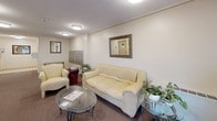 This single-floor lobby serves as the main communal entrance for a residential building, thoughtfully arranged to provide both functionality and a welcoming atmosphere. The space is finished with warm neutral tones throughout, featuring beige walls and patterned carpet that extends across the primary areas, while tiled flooring marks the entryway near the glazed exterior door.

The seating area forms the focal point of the lobby, furnished with a cream leather sofa and matching armchair arranged around a glass-top coffee table. Potted greenery and framed decorative mirrors on the walls add warmth and character to the space. Flush-mount ceiling light fixtures provide consistent, even illumination throughout.

Adjacent to the seating area, a bank of individual mailboxes lines one wall, accompanied by a glass-and-metal console table for residents' convenience. An elevator provides vertical access within the building, while a clearly marked exit leads toward the glazed entry vestibule, allowing natural light to filter into the space.

A dedicated office door, situated along one corridor, provides an on-site management presence. The hallway extends further into the building, maintaining the same neutral palette and carpet finish, creating a cohesive and tidy communal environment throughout.
