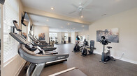 Located at 166 Caldy Knoll Dr in Sunbury, Ohio, this fitness center offers a generously sized, single-floor layout designed to accommodate a comprehensive range of workout activities. The space features a durable rubber-composite flooring that provides cushioning and traction throughout, making it well-suited for both cardio and strength training exercises.

The cardio section is prominently outfitted with multiple Spirit-brand treadmills arranged in a row, complemented by elliptical trainers and stationary bikes. Flat-screen televisions are mounted at eye level along the treadmill row, enhancing the workout experience. A dedicated strength training area includes a cable/multi-functional machine, a rack of dumbbells, and a weight bench, providing options for a full-body workout routine.

The space benefits from abundant natural light streaming through multiple large windows fitted with blinds, creating a bright and motivating atmosphere. Ceiling fans with recessed lighting maintain air circulation and comfort throughout. Large mirrors are positioned along one wall, expanding the perceived depth of the room. Motivational fitness artwork decorates the neutral-toned walls, contributing to an energetic yet clean aesthetic. The open floor plan allows for ample movement between equipment stations.