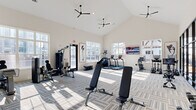 Located at 1994 US-72 E in Huntsville, Alabama, this contemporary fitness center is designed to provide a bright, energizing environment for a comprehensive workout experience. The space is defined by soaring vaulted ceilings fitted with sleek ceiling fans, which contribute to both the airy atmosphere and effective air circulation throughout. Large windows line the walls, flooding the interior with natural light and offering views of the surrounding outdoor amenity areas, including a resort-style pool and lounge space visible through dramatic floor-to-ceiling steel-framed glass panels.

The main workout area features patterned carpet tile flooring and houses an impressive array of equipment, including treadmills, ellipticals, stationary bikes, a rowing machine, a multi-function cable machine, and adjustable weight benches. A dedicated dumbbell rack provides a full range of free weights for strength training. A separate studio space is enclosed by sleek steel-framed glass partitions and features smooth hardwood-style flooring, making it ideal for yoga, stretching, or group fitness classes. This studio area is further enhanced by a large-scale abstract wall mural that adds a bold artistic element to the space.

Wall-mounted TVs, full-length mirrors, and curated artwork complete the modern aesthetic, creating a fitness facility that is both functional and visually refined.