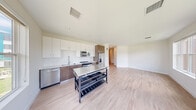 This contemporary single-floor unit showcases a clean, modern design highlighted by light hardwood flooring and crisp white walls throughout. A welcoming hallway leads into the heart of the home, where an open-concept kitchen and living area take center stage. The well-appointed kitchen features a two-tone cabinetry scheme pairing white upper cabinets with warm wood-toned lower cabinets, a white subway tile backsplash, stainless steel appliances including a refrigerator with ice maker, electric range, built-in microwave, and dishwasher. A moveable kitchen island with a stainless steel top adds functional workspace and flexibility. Large windows in the living and dining area flood the space with natural light and offer views of the surrounding neighborhood and green spaces. The bedroom is generously proportioned, with a sliding-door closet and a large window framing outdoor views. The bathroom is finished with a rich dark wood vanity, white countertop, large mirror with modern lighting, and a full tub and shower combination enclosed with classic white square tile. Recessed lighting and clean white baseboards complete the polished, move-in-ready aesthetic throughout the unit.