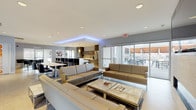 This contemporary single-floor space is thoughtfully designed as an open-plan communal area that seamlessly integrates multiple functional zones. The layout encompasses a well-appointed kitchen, relaxed lounge areas, a dining section, and direct access to an outdoor patio.

The kitchen is a standout feature, outfitted with stainless steel appliances including a full-size refrigerator, double wall ovens, and a dishwasher. Dark wood cabinetry lines the walls, complemented by a generous white-topped island with bar seating for several guests. Cylindrical pendant lights hang above, adding warmth to the space.

The lounge area is furnished with multiple contemporary sofas in neutral tan and cream tones arranged around a marble-topped coffee table. A linear fireplace set within a textured tile surround anchors one wall, with a wall-mounted television above it. Ambient blue LED cove lighting accents the tray ceiling, adding a modern atmosphere to the space.

Large sliding glass doors flood the interior with natural light and open directly onto an outdoor patio furnished with tables and chairs. Bold, colorful artwork displayed on the walls adds visual interest throughout. Large-format tile flooring runs throughout, unifying the entire space with a clean, polished aesthetic.