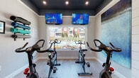 This spin studio is thoughtfully designed to deliver an engaging and motivating indoor cycling experience. The room is arranged with multiple stationary bikes positioned to face a large picture window, offering views of the surrounding exterior grounds and filling the space with natural light. Two wall-mounted flat-screen TVs are positioned above the window, ensuring every rider has a clear sightline for entertainment or instructional content. A third TV is mounted on a shiplap accent wall at the rear of the room, creating a focal point that adds architectural character and texture to the space.

The ceiling is finished in a deep charcoal tone, providing a striking contrast to the light-colored luxury vinyl plank flooring and bright white trim work throughout. Recessed lighting and a decorative multi-blade ceiling fan with integrated lighting ensure the room is well-lit and comfortable. A wall-mounted climate control unit supports temperature regulation during intense workouts.

Large-scale abstract artwork in oceanic blues and teals adorns the walls, contributing to a calm yet energizing ambiance. French doors with full-length glass panels provide access to an adjacent tiled space, adding connectivity and an open feel. The overall design balances functional fitness utility with polished, modern interior finishes.