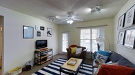 This single-floor space offers a comfortable and functional layout centered around an inviting living room. The room is anchored by a ceiling fan with integrated lighting, complemented by additional track lighting that ensures the space is well-lit throughout the day and evening. Large windows dressed with white plantation-style blinds allow generous natural light to fill the room, while a glass-paneled door provides direct access to an outdoor area, further enhancing the sense of openness. The living room features warm, carpeted flooring and is furnished with a plush brown sofa accented with colorful throw pillows, creating a relaxed and welcoming atmosphere. A bold navy and white striped area rug defines the seating area and adds a graphic, modern touch. An open media console in a matte black finish houses the flat-screen television, while framed wall art arranged in a gallery-style grouping adds personality and visual interest to the walls. The textured ceiling adds a classic residential character to the overall aesthetic. The space strikes a balance between casual comfort and thoughtful design, making it a practical and livable environment.