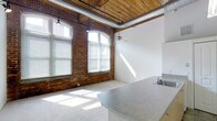 This single-floor loft space showcases a compelling blend of industrial and residential design elements that work together to create a distinctive, character-rich environment. The open living and kitchen area serves as the heart of the space, where original exposed brick walls rise to meet a rustic wood-beam ceiling with visible ductwork — hallmarks of thoughtful adaptive reuse architecture. Large arched windows line the brick wall, inviting an abundance of natural light that spills across the floors throughout the day. A ceiling fan provides comfort and adds to the airy feel of this generously proportioned main room. The kitchen features a curved countertop with a stainless steel sink, providing a functional and stylish workspace that integrates seamlessly into the open-plan layout. A hallway with soft carpet underfoot connects the living area to additional rooms, lined with the same white walls and accented overhead by exposed wood ceiling planks and industrial pendant lighting. A classic paneled white door with brushed nickel hardware adds a clean, refined contrast to the raw industrial backdrop. Throughout the space, the interplay of reclaimed materials, natural light, and clean white surfaces creates an inviting and versatile atmosphere.