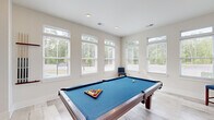Located at 1005 Sonoran Cir in Summerville, SC, this single-floor recreational space is thoughtfully divided into two functional areas designed for leisure and entertainment. The layout flows seamlessly between a dedicated billiards room and a cozy lounge area, both finished in a clean, modern aesthetic with neutral walls and light-toned luxury vinyl plank flooring throughout.

The billiards room is the centerpiece of the space, anchored by a full-size pool table with a vibrant blue felt surface. Large double-hung windows with white blinds line the walls, flooding the room with natural light and offering views of the surrounding greenery. Recessed lighting supplements the natural light, while a wall-mounted cue rack keeps equipment neatly organized. The shiplap accent wall adds a subtle coastal charm to the space.

The adjoining lounge area provides a relaxed setting for socializing, featuring plush upholstered armchairs, a wooden coffee table atop a patterned area rug, and a wall-mounted flat-screen television. A multi-tiered open shelving unit displays an assortment of board games, making this area ideal for group entertainment. Coastal-themed wall art ties the two areas together, creating a cohesive and inviting atmosphere throughout the space.