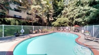 This inviting outdoor pool area offers a tranquil retreat set against a lush backdrop of mature evergreen and deciduous trees. The generously sized, kidney-shaped pool features clear turquoise water and is framed by a classic brick coping detail, adding a timeless aesthetic to the space. The surrounding deck is finished with a textured aggregate surface that provides both durability and slip resistance, and is equipped with multiple lounge chairs and seating arrangements—including a shaded table and umbrella area—ideal for relaxing in the sun or gathering with others. A white metal perimeter fence encloses the pool area, with a gated entry point providing controlled access. Adjacent to the pool is a pool house structure finished in dark wood siding, housing restroom and shower facilities, with additional seating positioned just outside its entrance. Potted flowers and plantings add a welcoming, decorative touch throughout the deck. The towering trees surrounding the space provide natural shade and privacy, creating a peaceful, park-like atmosphere. The multi-story residential building visible in the background, with its wood-paneled balconies and landscaped hillside, underscores the well-maintained character of the overall community this amenity serves.