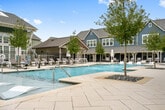 Located in Birmingham, Alabama, this resort-style pool complex offers a richly appointed outdoor amenity experience designed for relaxation and recreation. The centerpiece is a generously sized, crystal-clear swimming pool divided into distinct sections, including a sun shelf with in-water chaise loungers ideal for wading and sunbathing. Water features add a dynamic element to the pool's surface, enhancing the resort ambiance throughout the space.

The expansive pool deck is finished with elegant herringbone-patterned stone pavers and is thoughtfully landscaped with young trees planted in decorative rock beds, providing natural accents across the hardscape. Rows of cushioned lounge chairs and market umbrellas line the perimeter, offering ample space for sunbathing and relaxation.

A covered outdoor pavilion area features a pool table and additional seating, providing a shaded gathering spot for social activities. The surrounding architecture includes a well-appointed clubhouse building with a standing-seam metal roof and large glass doors, alongside charming residential-style structures clad in green and blue board-and-batten siding that frame the complex.

The entire area is enclosed by black iron fencing, ensuring privacy and security while maintaining an open, airy feel. Together, these elements create a cohesive and inviting outdoor retreat.