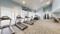 Located at 500 Waters Edge Dr in Newport News, VA, this fitness center offers a comprehensive workout facility designed to support a full range of exercise routines. The space is composed of a generously sized main exercise room and a connecting hallway, together spanning a single floor.

The exercise room is the centerpiece of the facility, boasting soaring vaulted ceilings that create an open, airy atmosphere. Soft blue-gray walls complement the neutral-toned carpet flooring, lending a clean and energizing ambiance. Large windows with white blinds allow abundant natural light to fill the room, enhancing the sense of space. Pendant light fixtures hang from the ceiling, adding a polished finishing touch.

The room is fully equipped with multiple treadmills, elliptical machines, stationary spin bikes, stair climbers, a cable weight machine, a dumbbell rack with free weights, a flat bench, and stability balls — providing everything needed for both cardio and strength training. Bold motivational wall graphics, including "Don't Wish For It, Work For It," add an inspiring energy to the environment.

The hallway entrance features warm hardwood-style flooring, recessed lighting, a built-in water fountain, and frosted glass sliding panels that offer a stylish transition into the main workout area. A security camera ensures a safe environment throughout.